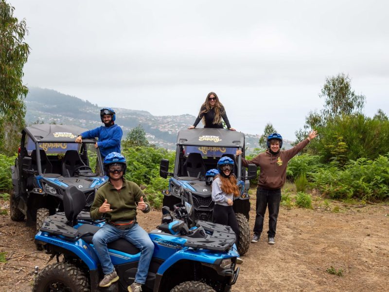 A group of six excited participants in helmets pose with thumbs up beside two blue Can-Am buggies from Let’s Buggy, overlooking misty hills and distant mountains in Madeira’s highlands, during a Let's Buggy's Laurissilva experience in Madeira.