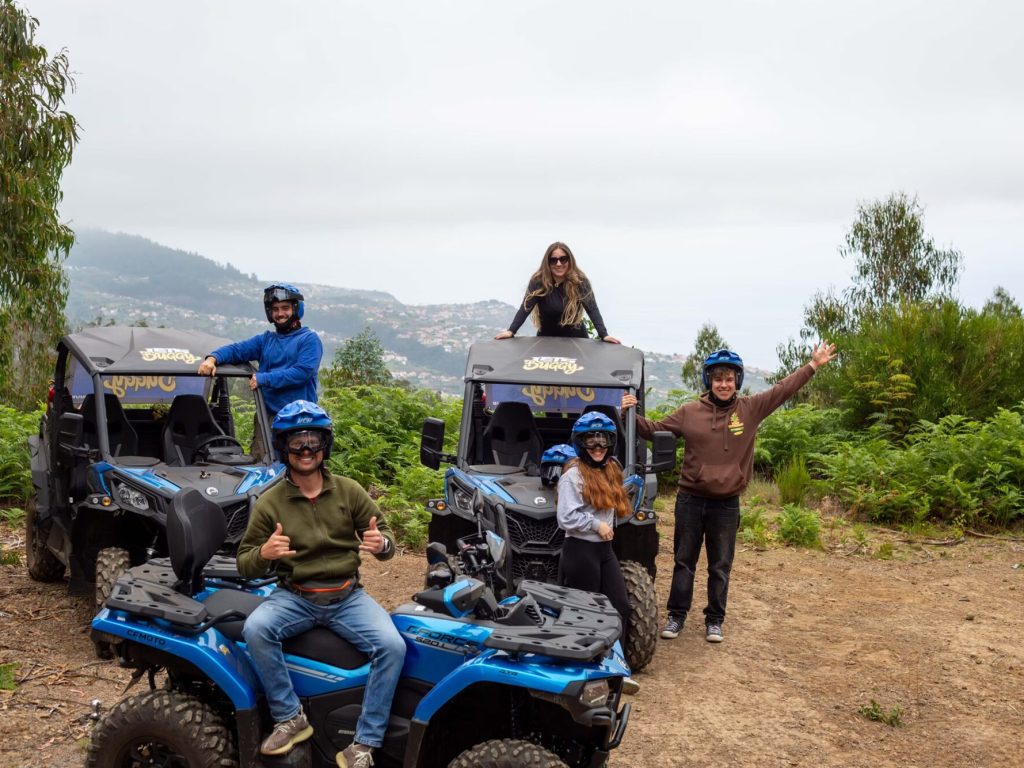 A group of six excited participants in helmets pose with thumbs up beside two blue Can-Am buggies from Let’s Buggy, overlooking misty hills and distant mountains in Madeira’s highlands, during a Let's Buggy's Laurissilva experience in Madeira.