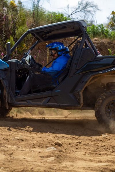 A driver in a blue helmet powers a Can-Am Maverick buggy through a dusty corner, kicking up a cloud of dirt on a rugged off-road track lined with wildflowers and greenery in Madeira Laurissilva forest, during a Let's Buggy's Laurissilva experience in Madeira.