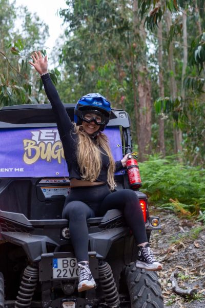 A woman in black sportswear and blue helmet waves enthusiastically from the back of a Let’s Buggy Can-Am Maverick amid dense Laurissilva forest greenery in Madeira, during a Let's Buggy's Laurissilva experience in Madeira.