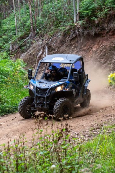 Two passengers smile excitedly in a black Can-Am buggy kicking up dust on a lush green off-road trail surrounded by trees and wildflowers during a Let's Buggy happy hour tour.
