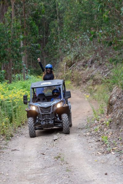 A blonde woman passenger in a blue helmet waves triumphantly from the back of a Let’s Buggy Can-Am buggy as it navigates a sunny dirt trail lined with yellow wildflowers and dense green forest in Madeira’s Laurissilva, during a Let's Buggy's Happy Hour Tour experience in Madeira.