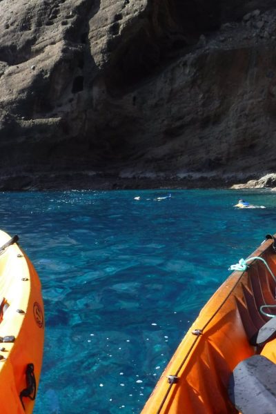 Garajau sea kayaking experience by Madeira Radical, with two orange kayaks floating on crystal-clear blue water beside dramatic rocky cliffs and snorkelers swimming in the marine reserve.