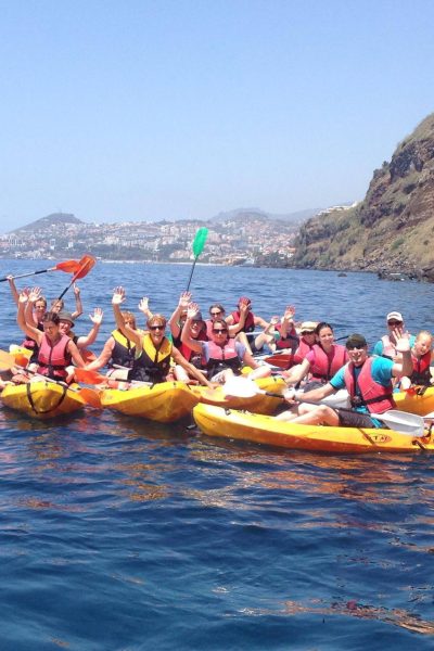 Large group of kayakers in bright yellow kayaks at Garajau, waving and smiling on the calm blue sea with Madeira’s coastal cliffs and Funchal skyline in the background, an experience by Madeira Radical.