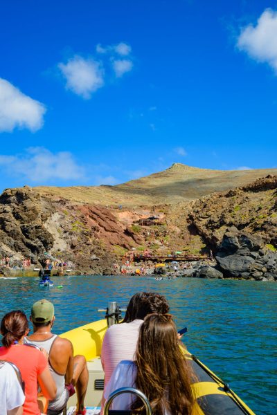 The sea, rocky land and blue cloudy sky on the horizon, and People on a Madeira Sea Emotions boat tour arriving at the Cais do Sardinha.