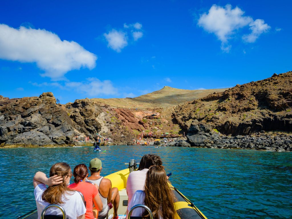The sea, rocky land and blue cloudy sky on the horizon, and People on a Madeira Sea Emotions boat tour arriving at the Cais do Sardinha.