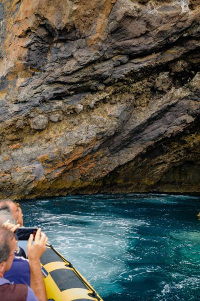 A yellow RIB boat packed with excited tourists heads into a dramatic volcanic sea cave with turquoise waters and orange cliffs ahead taking pictures with a a mobile phone, a boat tour experience by Madeira Sea Emotions.