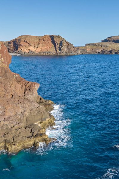 A boat leaves a white wake across blue waters off Ponta de São Lourenço's dramatic, sunlit volcanic cliffs and headlands, a boat tour experience by Madeira Sea Emotions.