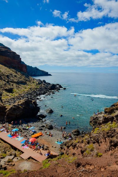 Cais do Sardinha with people relaxing taking sunbathing and swimming on the sea and a yellow tour boat of Madeira Sea Emotions leaving with people for another tour around the Madeira Coast.