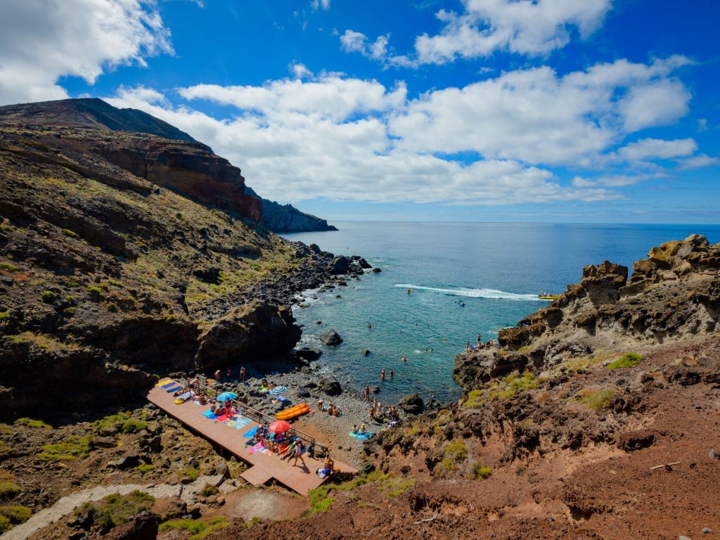 Cais do Sardinha with people relaxing taking sunbathing and swimming on the sea and a yellow tour boat of Madeira Sea Emotions leaving with people for another tour around the Madeira Coast.