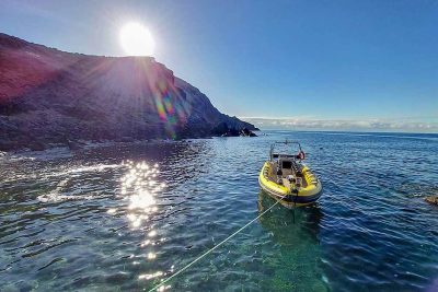 A yellow Madeira Sea Emotions RIB boat rests on crystal-clear water at Ponta de São Lourenço, with the sun setting behind rugged cliffs and casting rainbow flares across the calm cove.