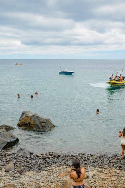 Crowds relax on the rocky shore and swim in the calm cove at Cais do Sardinha, while a yellow Madeira Sea Emotions tour boat is taking more people to do some touring on the coast.