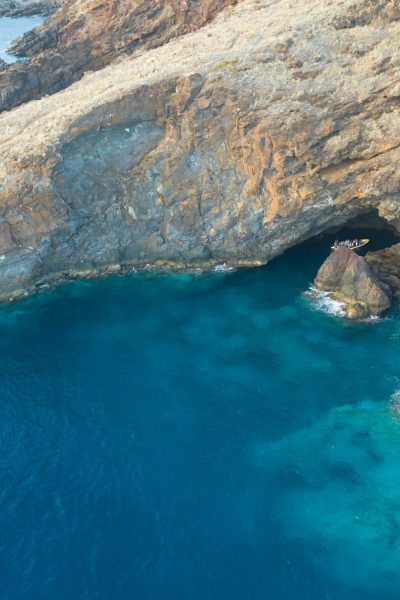 A rugged volcanic cove at Ponta de São Lourenço, where turquoise water laps against the cliffs and a yellow Madeira Sea Emotions tour boat rests at the entrance of a sea cave.