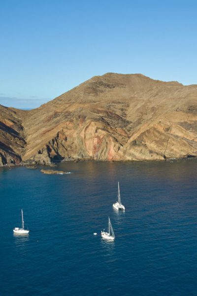 A tranquil bay at Ponta de São Lourenço, where sailboats anchor beneath layered golden and red volcanic cliffs rising steeply from the deep blue Atlantic.