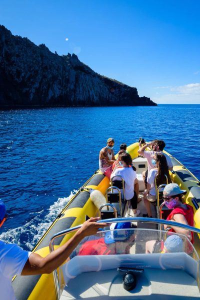 A yellow RIB boat full of tourists and a guide in a cap cruises past Ponta de São Lourenço's towering dark cliffs and a rock arch under bright blue skies, a boat tour experience by Madeira Sea Emotions.