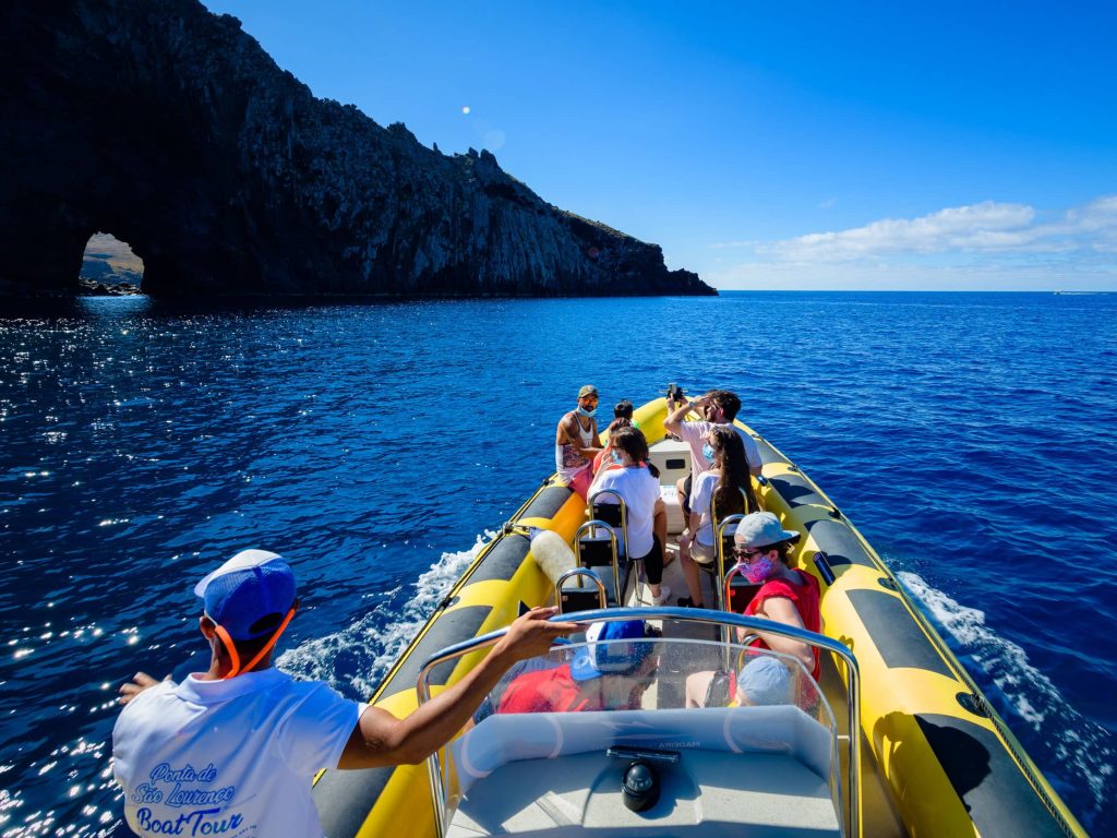 A yellow RIB boat full of tourists and a guide in a cap cruises past Ponta de São Lourenço's towering dark cliffs and a rock arch under bright blue skies, a boat tour experience by Madeira Sea Emotions.