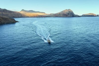 A boat leaves a white wake across blue waters off Ponta de São Lourenço's dramatic, sunlit volcanic cliffs and headlands, a boat tour experience by Madeira Sea Emotions.