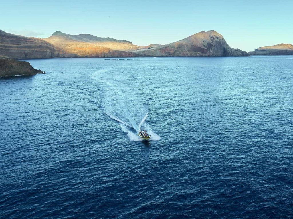 A boat leaves a white wake across blue waters off Ponta de São Lourenço's dramatic, sunlit volcanic cliffs and headlands, a boat tour experience by Madeira Sea Emotions.