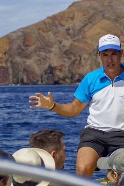 A guide on a Madeira Sea Emotions boat in the sea an with the Ponta de S.Lourenço in the background talking with clients about what they are seeing,a boat tour experience by Madeira Sea Emotions.