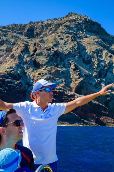 A guide in sunglasses and cap on a Madeira Sea Emotions boat spreads arms wide toward towering volcanic cliffs under sunny skies. a boat tour experience by Madeira Sea Emotions.