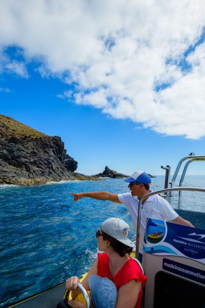 A guide in a cap points from a blue Madeira Sea Emotions RIB boat toward a path on volcanic cliffs under partly cloudy skies, a boat tour experience by Madeira Sea Emotions.
