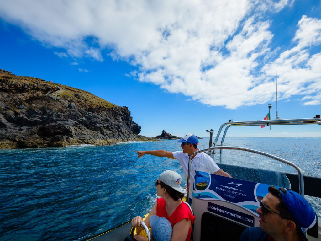 A guide in a cap points from a blue Madeira Sea Emotions RIB boat toward a path on volcanic cliffs under partly cloudy skies, a boat tour experience by Madeira Sea Emotions.