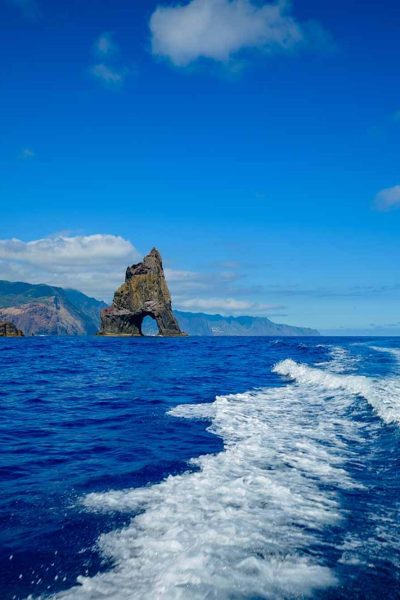A boat leaves a frothy wake as it passes rugged volcanic cliffs and a tall rock pillar with an arch under clear blue skies, a boat tour experience by Madeira Sea Emotions.