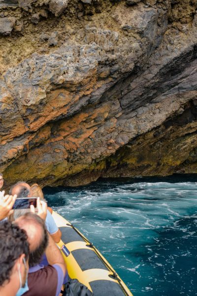 A yellow RIB boat packed with excited tourists heads into a dramatic volcanic sea cave with turquoise waters and orange cliffs ahead taking pictures with a a mobile phone, a boat tour experience by Madeira Sea Emotions.