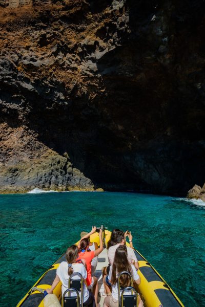 A yellow RIB boat packed with excited tourists heads into a dramatic volcanic sea cave with turquoise waters and orange cliffs ahead., a boat tour experience by Madeira Sea Emotions.