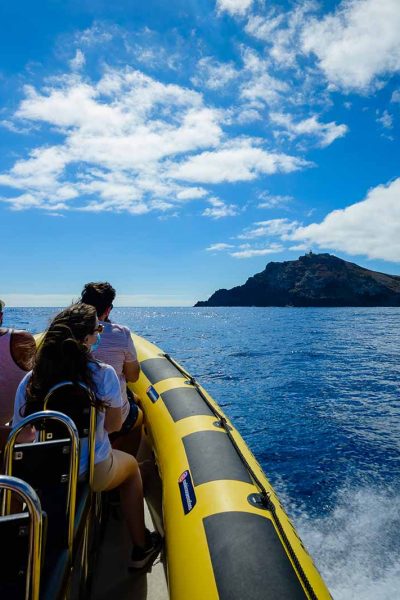 Tourists in colorful clothes ride a yellow RIB boat toward a rugged volcanic islet under partly cloudy blue skies, a boat tour experience by Madeira Sea Emotions.