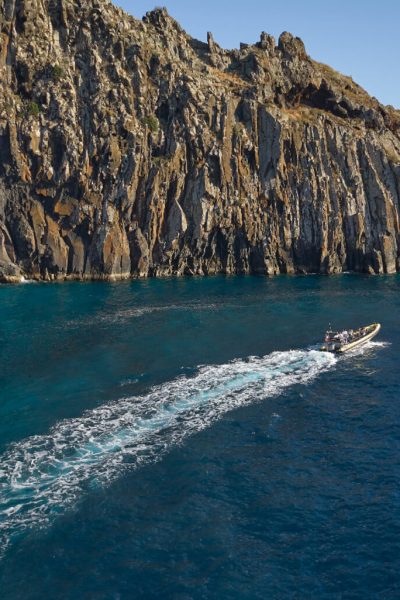 A boat leaves a white wake across blue waters off Ponta de São Lourenço's dramatic, sunlit volcanic cliffs and headlands, a boat tour experience by Madeira Sea Emotions.
