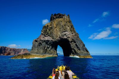 Tourists on a yellow RIB boat approach a massive volcanic rock formation with a dramatic archway rising from the deep blue ocean, a boat tour experience by Madeira Sea Emotions.