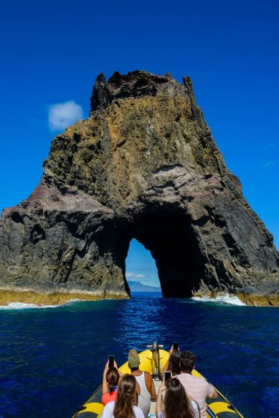 Tourists on a yellow RIB boat approach a massive volcanic rock formation with a dramatic archway rising from the deep blue ocean, a boat tour experience by Madeira Sea Emotions.