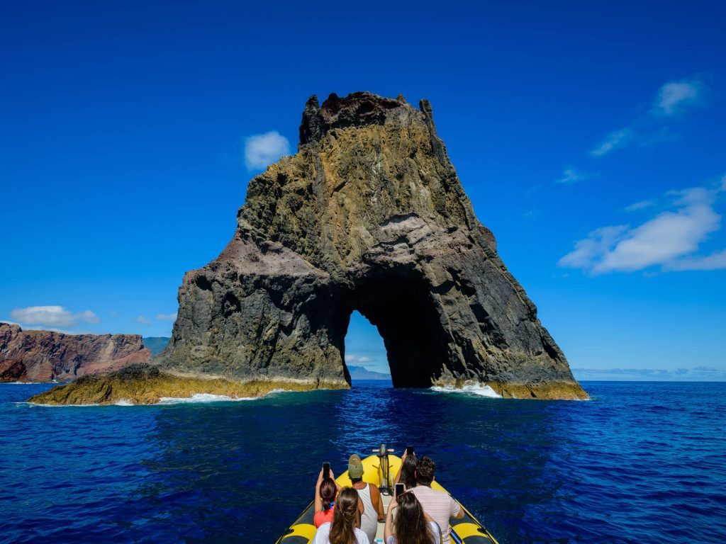 Tourists on a yellow RIB boat approach a massive volcanic rock formation with a dramatic archway rising from the deep blue ocean, a boat tour experience by Madeira Sea Emotions.