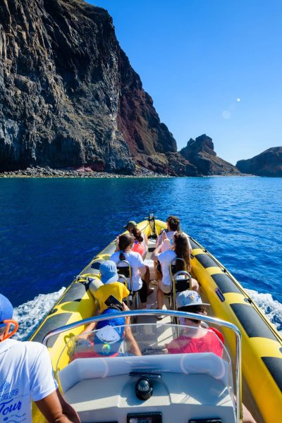 A yellow RIB boat full of tourists and a guide in a cap cruises past Ponta de São Lourenço's towering dark cliffs and a rock arch under bright blue skies, a boat tour experience by Madeira Sea Emotions.