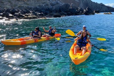 Two orange kayaks paddle along Ponta de São Lourenço's dark volcanic coastline, with life-vested explorers enjoying the turquoise waters and sunny skies, a Kayaking experience by Madeira Sea Emotions.