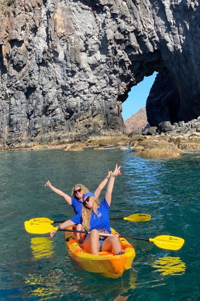 2 girls Kayaking in a full day pack of fun along Ponta de São Lourenço's rugged volcanic coast, framed by a massive rock archway in a kayaking Madeira Sea Emotions experience.