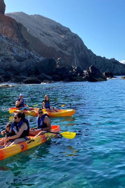 Two orange kayaks paddle along Ponta de São Lourenço's dark volcanic coastline, with life-vested explorers enjoying the turquoise waters and sunny skies, a Kayaking experience by Madeira Sea Emotions.
