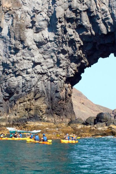 Kayakers in yellow and orange boats paddle through turquoise waters along Ponta de São Lourenço's rugged volcanic coast, framed by a massive rock archway in a kayaking Madeira Sea Emotions experience.