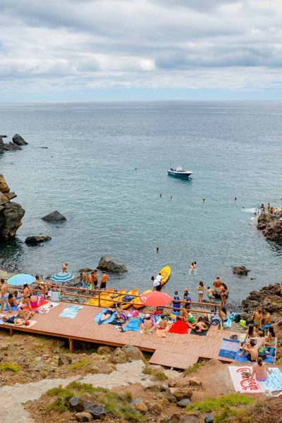 Cais do Sardinha with people relaxing taking sunbathing and swimming on the sea and a yellow tour boat of Madeira Sea Emotions leaving with people for another tour around the Madeira Coast.