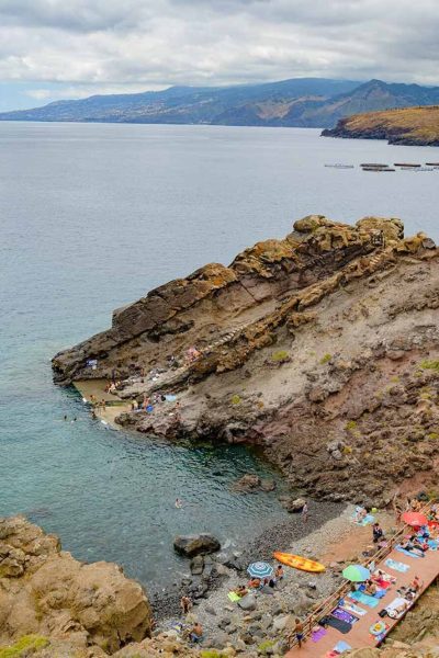 Cais do Sardinha filled with people relaxing, swimming and kayaking, under a cloudy grey sky, and Funchal coast on the horizon.