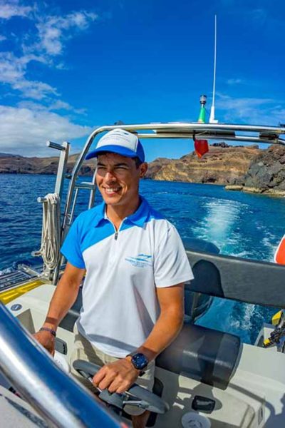 Boat skipper at the helm of a yellow speedboat off Ponta de São Lourenço, with rugged cliffs and deep blue sea in the background, during a boat tour experience by Madeira Sea Emotions.