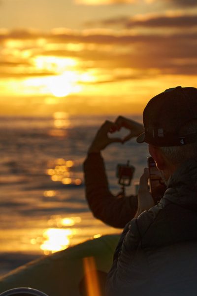 two people on a boat one doing a shape heart with hands and another filming it with a mobile phone with a sunset over the sea in the back during a boat tour experience by Madeira Sea Emotions.