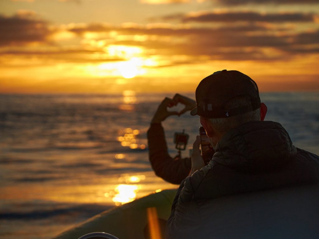 two people on a boat one doing a shape heart with hands and another filming it with a mobile phone with a sunset over the sea in the back during a boat tour experience by Madeira Sea Emotions.