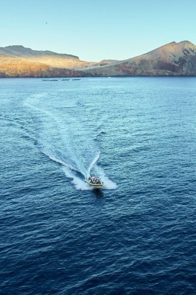 A boat leaves a white wake across blue waters off Ponta de São Lourenço's dramatic, sunlit volcanic cliffs and headlands, a boat tour experience by Madeira Sea Emotions.