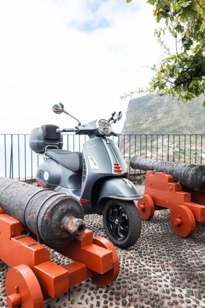 A grey Vespa scooter is parked on a stone terrace between three old iron cannons on wooden mounts, overlooking dramatic coastal cliffs and the distant village below in Nuns Valley, Madeira. The vehicle is a Vespa Primavera/GTS 125cc model, the type offered for rent to explore the island’s winding mountain roads and scenic viewpoints by Vespa Madeira Scooter Rent.