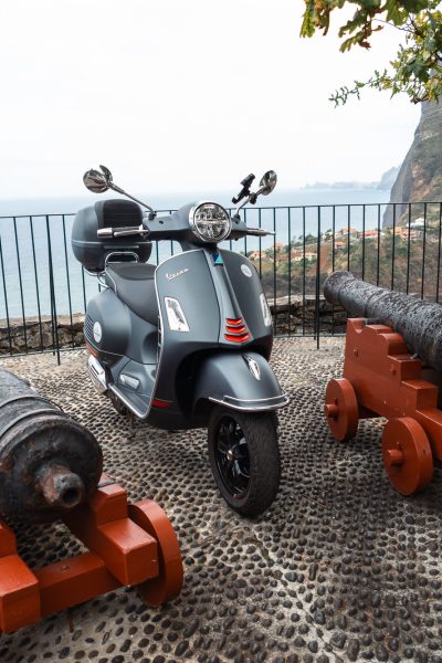 A modern grey Vespa scooter is parked on a stone terrace between three old iron cannons on wooden mounts, overlooking dramatic coastal cliffs and the distant village below in Nuns Valley, Madeira. The vehicle is a Vespa Primavera/GTS 125cc model, the type offered for rent to explore the island’s winding mountain roads and scenic viewpoints by Vespa Madeira Scooter Rent.