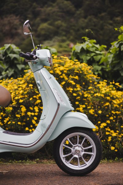 Side view close up of a green Vespa parked in a road with yellow flowers on the background and some green fields somewhere in Madeira.The type of scooter offered for rent to explore the Madeira island’s winding mountain roads and scenic viewpoints by Vespa Madeira Scooter Rent.