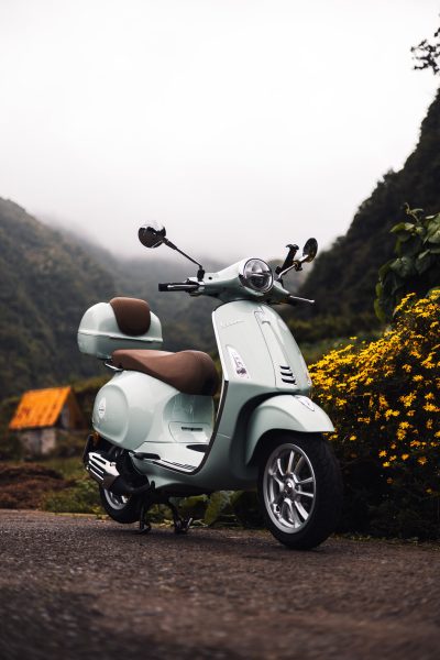 A green Vespa parked in a road with fog and a grey sky on the back, yellow flowers on the right and on the background a palheiro house on a Moutain with green fields somewhere in Madeira.The type of scooter offered for rent to explore the Madeira island’s winding mountain roads and scenic viewpoints by Vespa Madeira Scooter Rent.