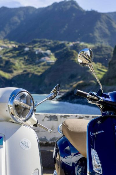 A close up of Two Vespa scooters,a blue one and a white one by Vespa Madeira Scooter Rent parked and on the back the Sea and mountains of Madeira Island. These are the type of scooters offered for rent to explore the Madeira island’s winding mountain roads and scenic viewpoints by Vespa Madeira Scooter Rent.